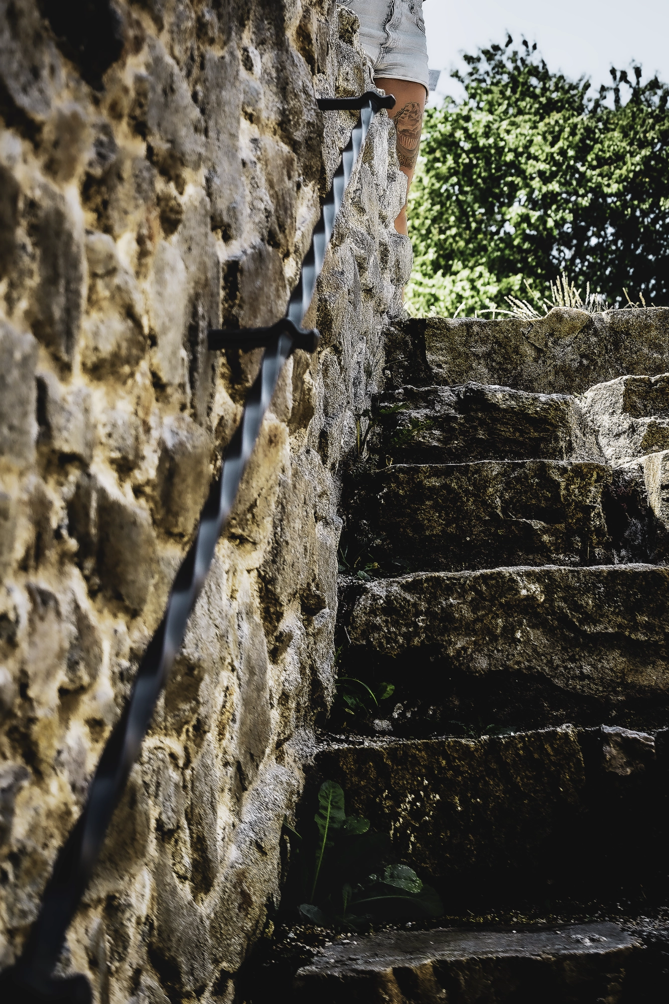 Rough stone steps and an iron handrail rising toward a partially visible tattooed figure in sunlight.