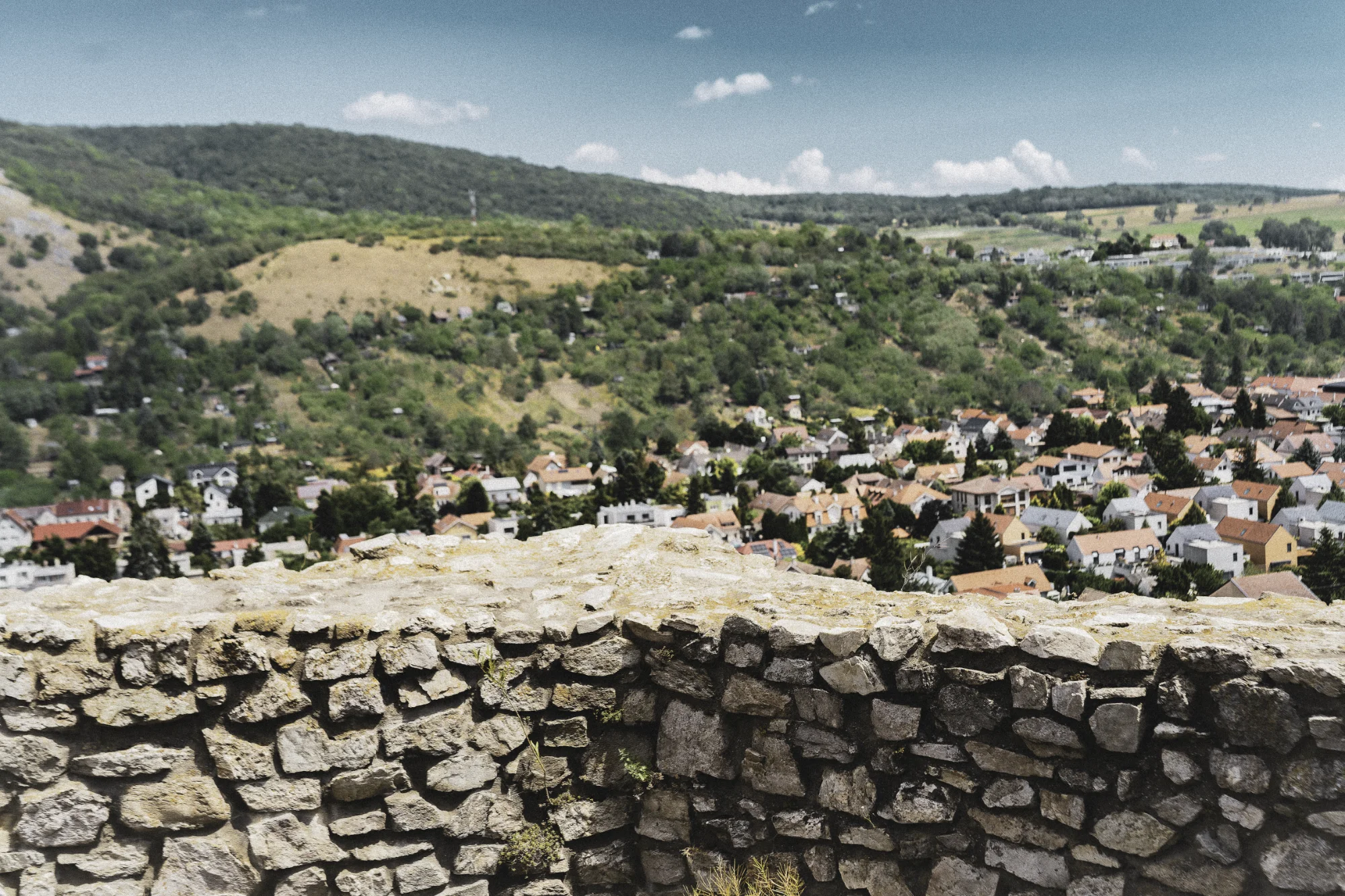 Rough stone parapet overlooking a town of red roofs among green hills beneath a blue sky.