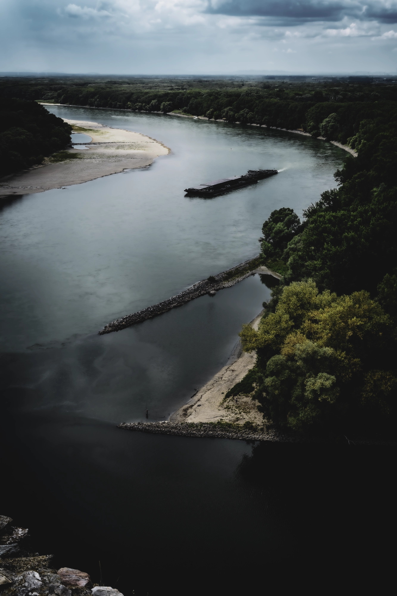 Cargo vessel moving along the Danube below Devín Castle, bordered by sandbanks and forest under dark clouds.