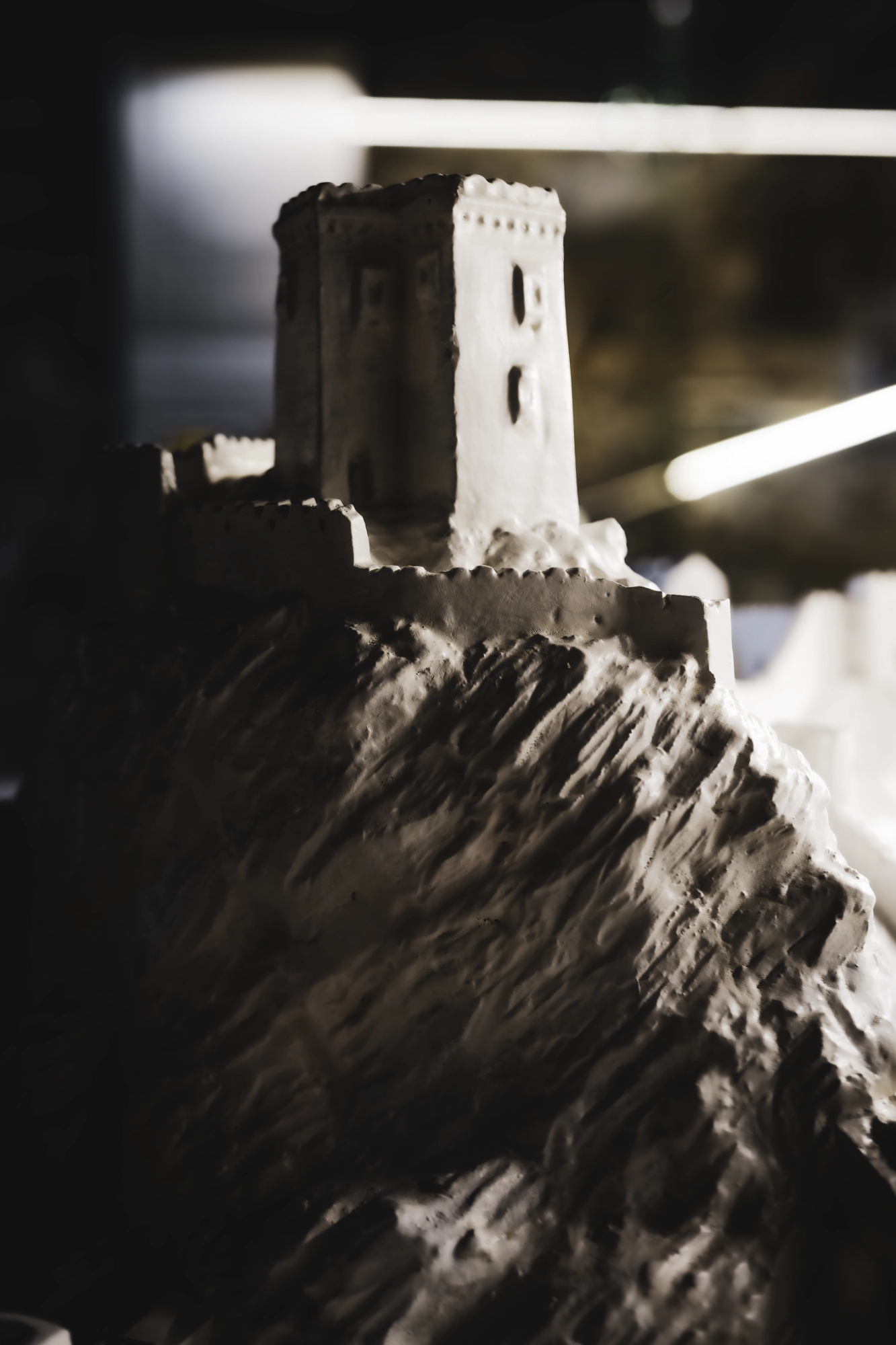 Small pale fortress model on textured terrain, dramatically lit against a dark blurred background.