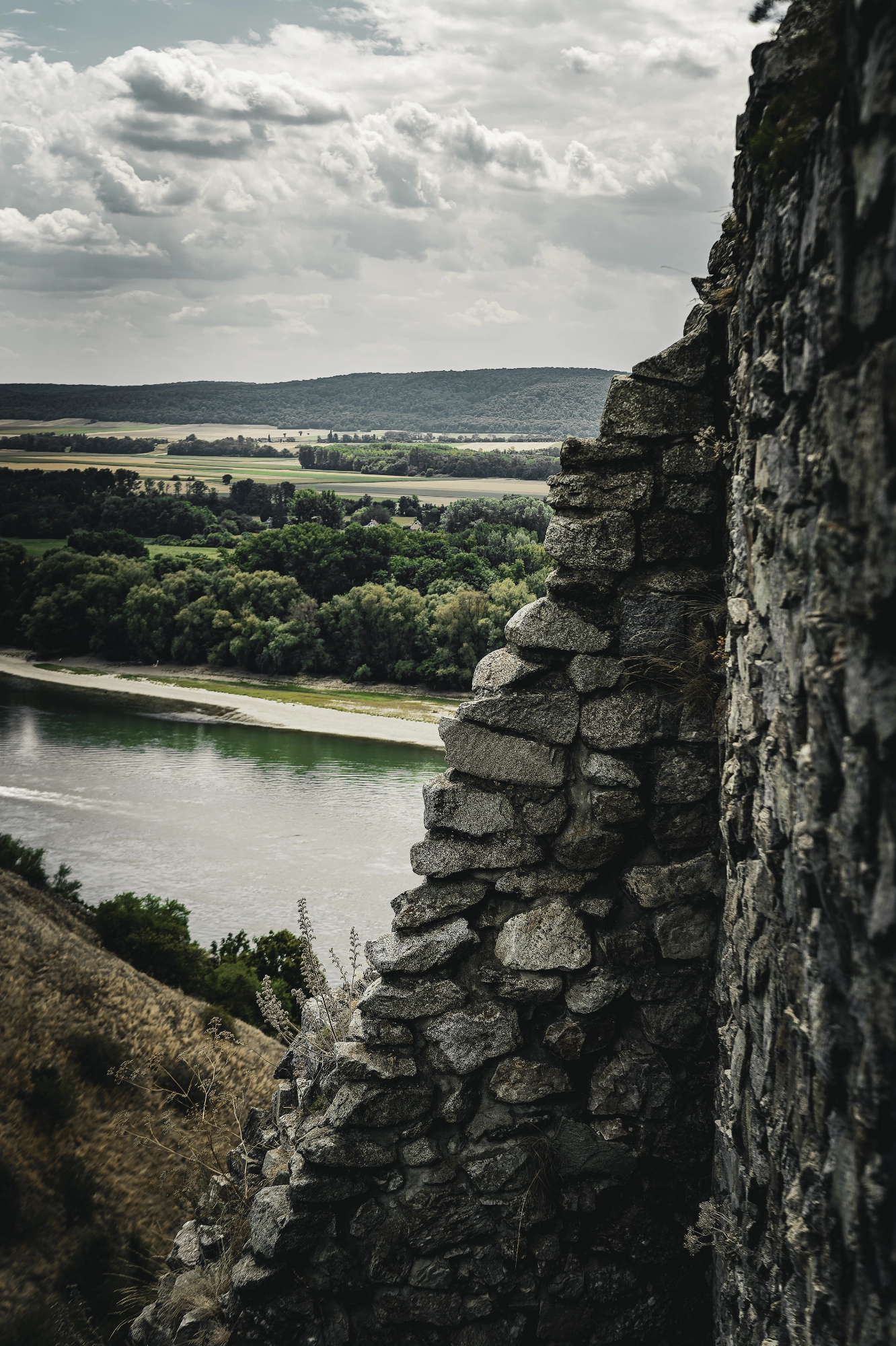 Broken stone wall framing a river, green fields and wooded hills beneath layered clouds.