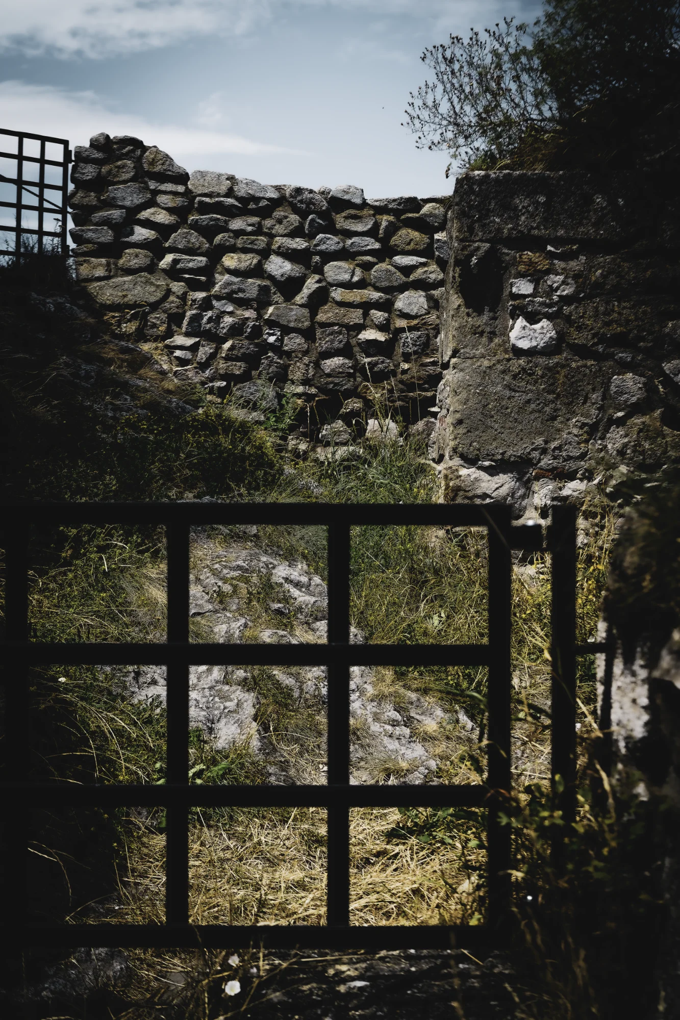 Dark iron grid in front of sunlit grass and weathered stone walls beneath a pale blue sky.