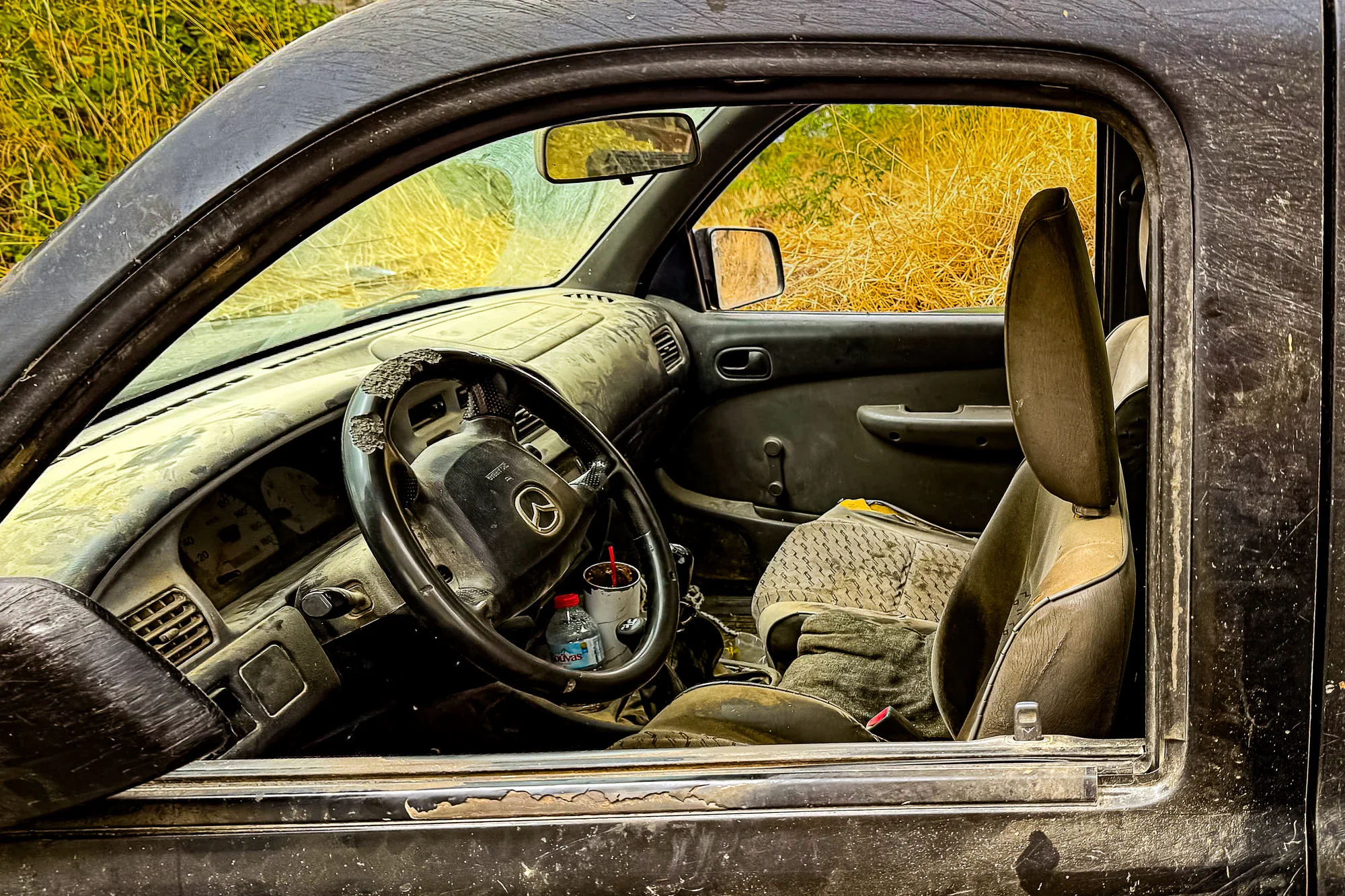 Dusty interior of an older Mazda with a worn steering wheel, open side window and dry Cretan grass beyond.