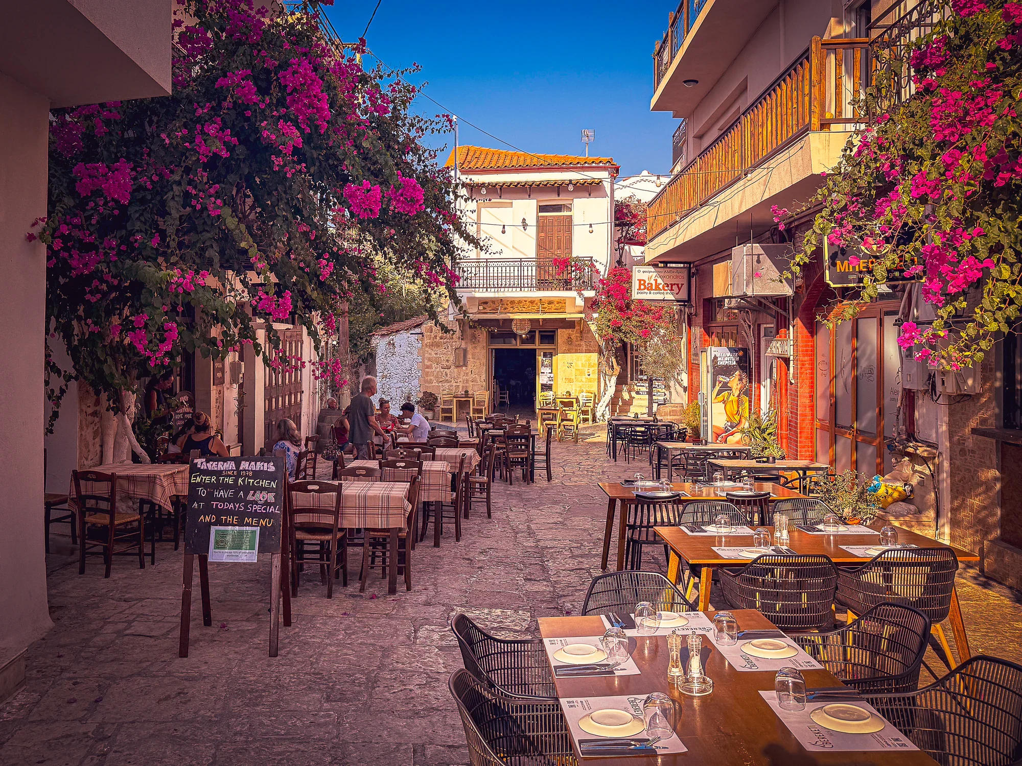 Cobbled lane in Panormos lined with bougainvillea, restaurant tables and several seated diners in evening light.