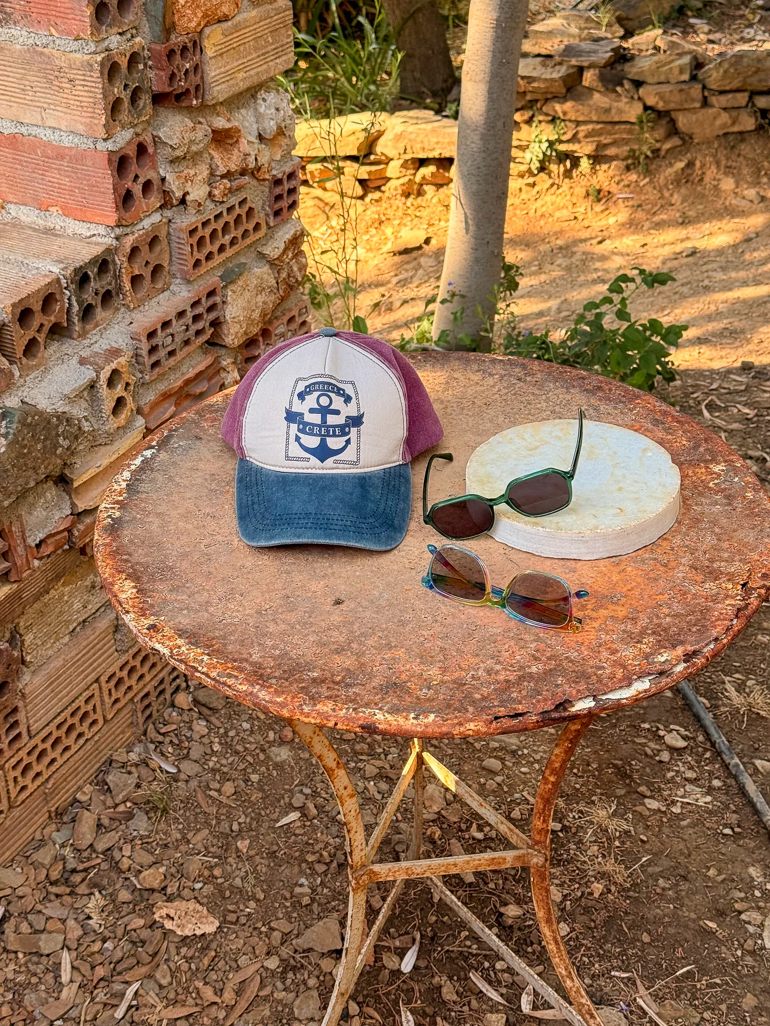 Two pairs of sunglasses and a Crete cap resting on a rusted round table against rough brick and earth.