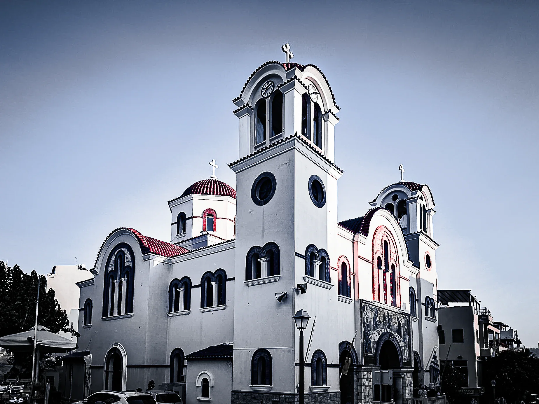 White Greek Orthodox church with belfries, a clock tower, crosses and red roof accents against a pale Cretan sky.