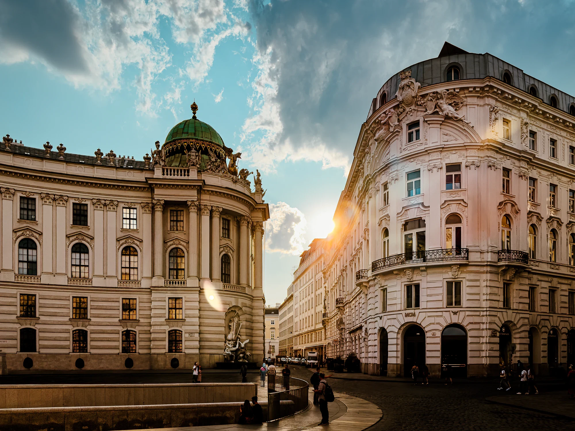 Late sunlight breaking between ornate facades at Vienna’s Michaelerplatz, with the Hofburg dome and people below.