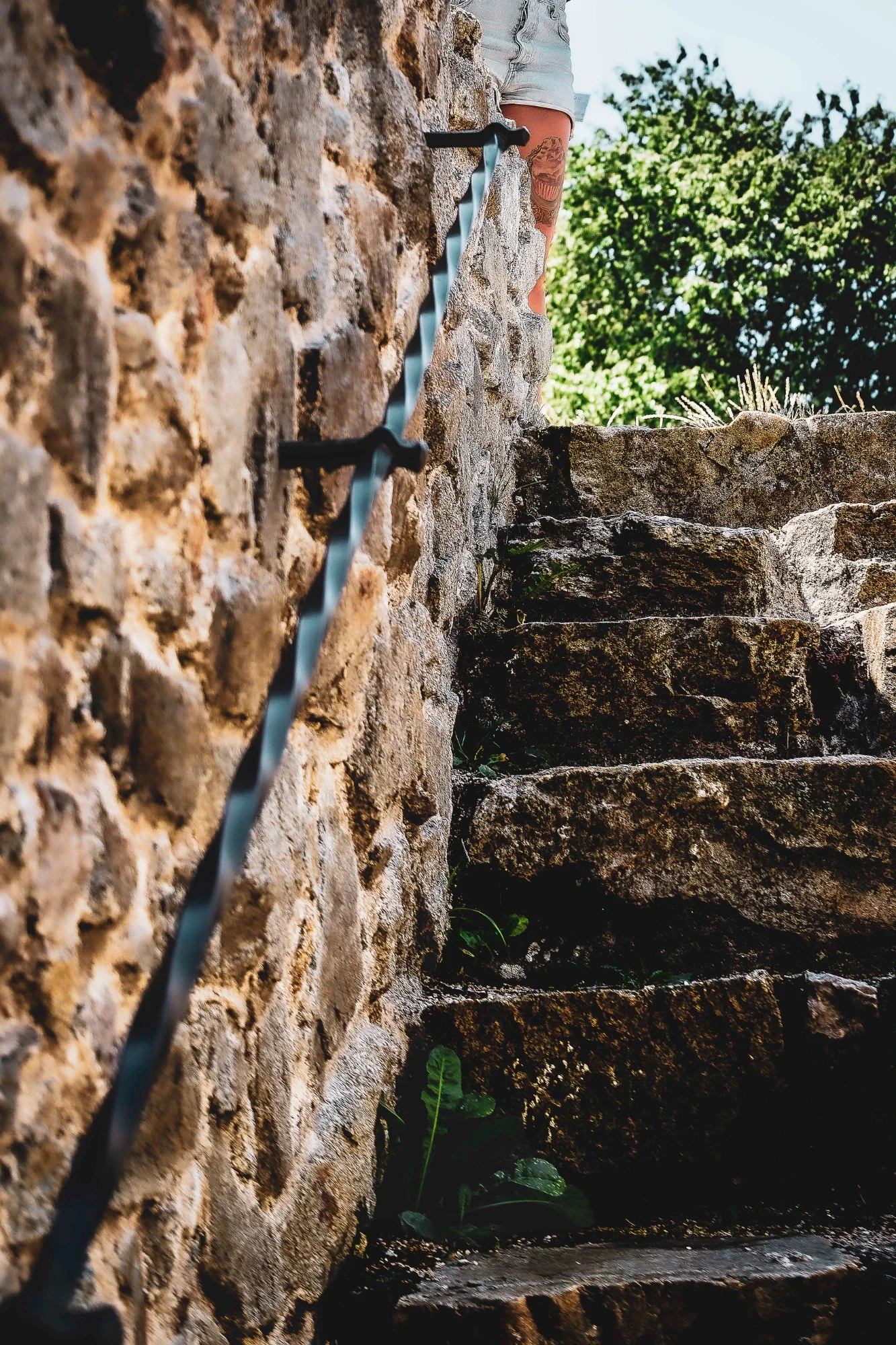 Rough stone steps and a curved iron handrail rising toward a cropped tattooed leg in sunlight.
