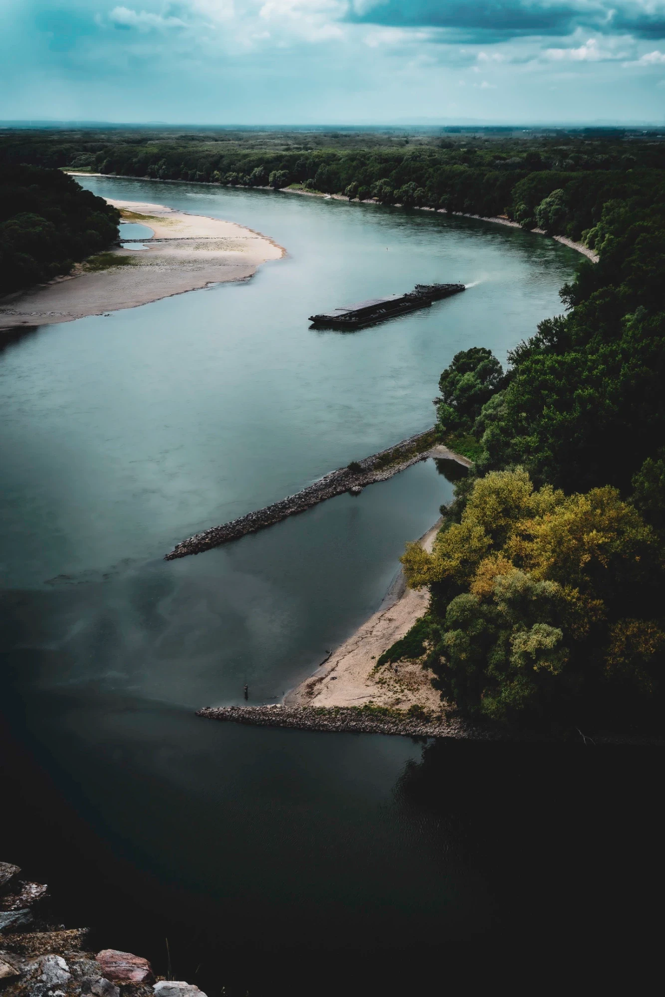 Cargo barge moving along the Danube between pale riverbanks and forest under low dark clouds.