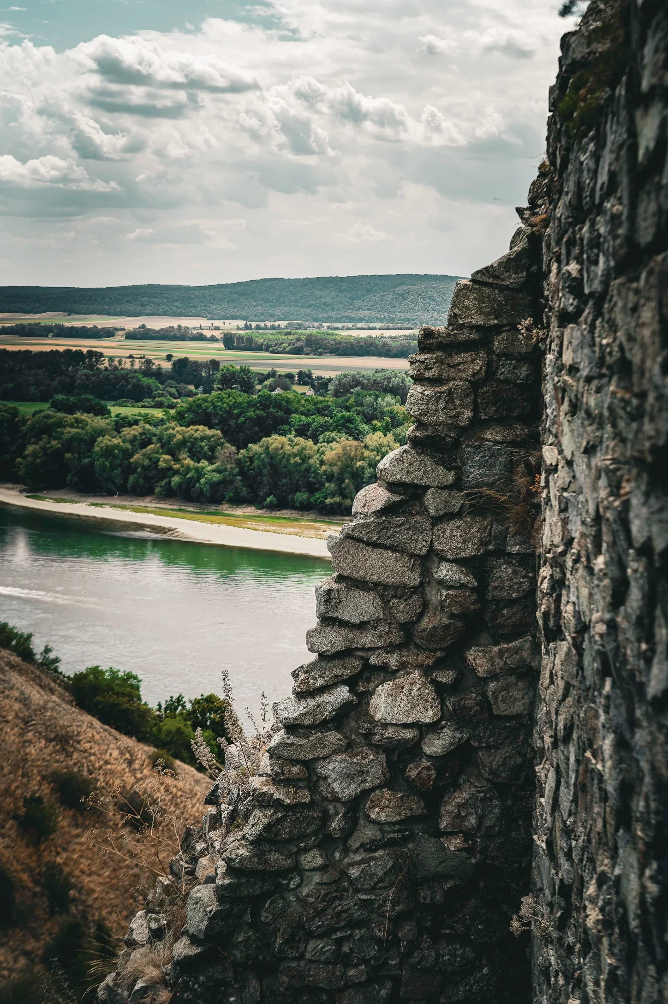 Broken stone wall framing a river, green fields and wooded hills beneath layered clouds.
