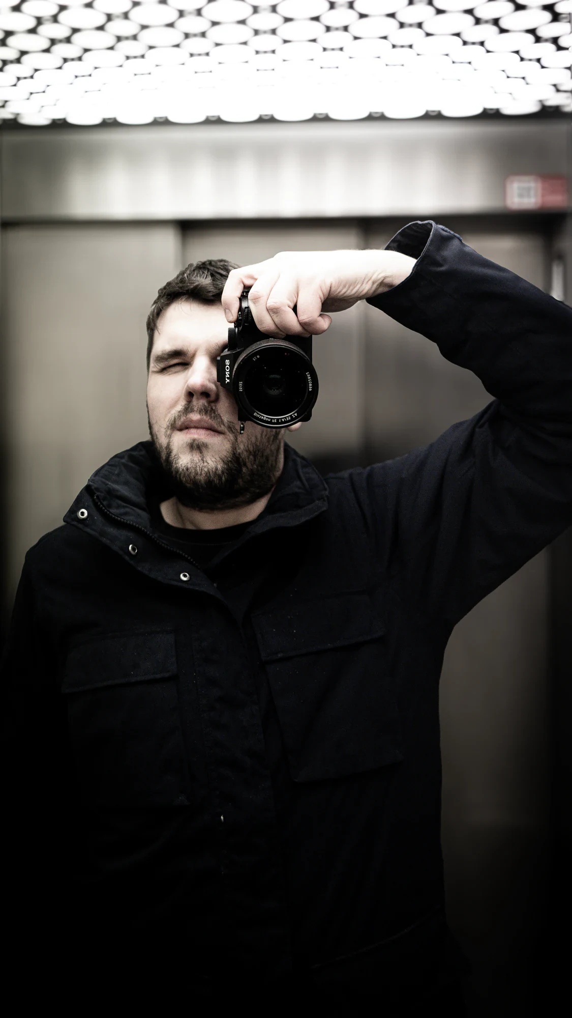 Simon Simons holding a Sony camera to his eye in a mirrored lift beneath a grid of circular lights.